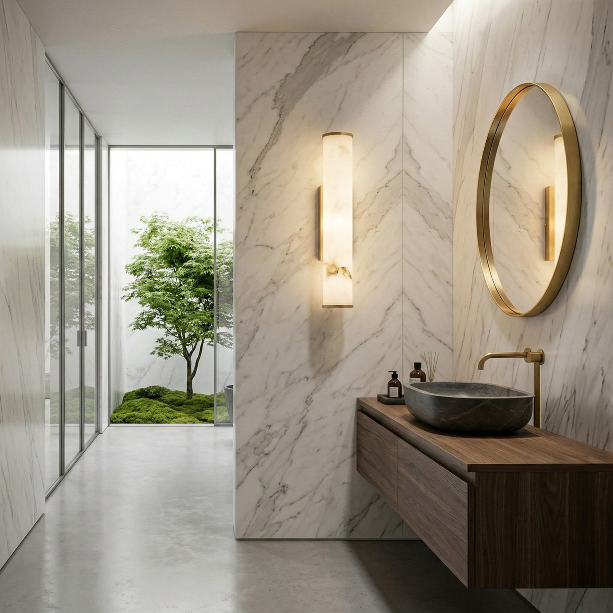 Modern bathroom featuring marble walls, a round gold-framed mirror, stone vessel sink on wooden vanity, gold faucet, Montavo LED Large Tube Alabaster Wall Light in soft white & brushed brass, and a window view of mossy greenery.