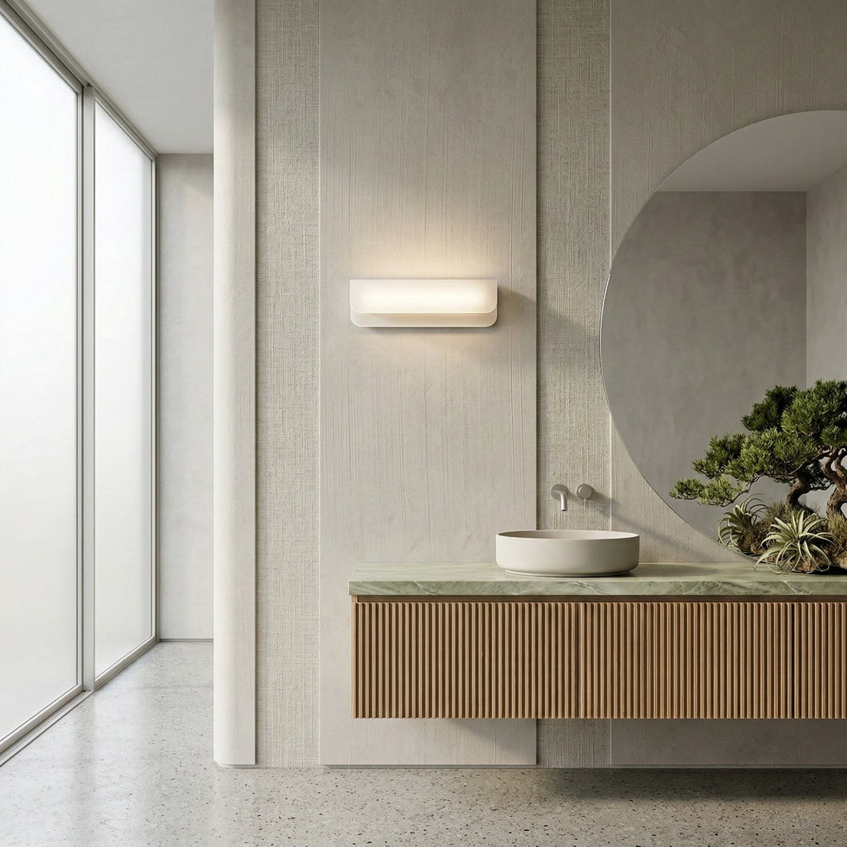 Minimalist bathroom with a large round mirror, the Velisse LED Horizontal Alabaster Wall Light in soft white, a floating vanity with ribbed wood front, round vessel sink, small bonsai tree, and a frosted glass window.