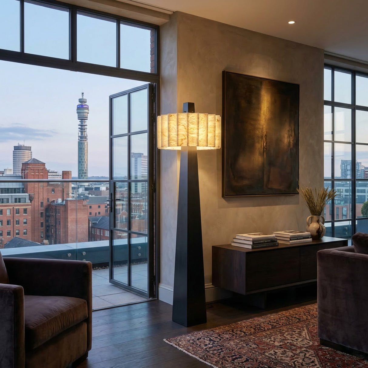 A modern living room with large windows and a balcony overlooks the city, including the BT Tower. The Venus 1 Light Alabaster Floor Lamp in Soft White & Matte Black glows by a dark console, complemented by a rug and abstract art for cozy elegance.