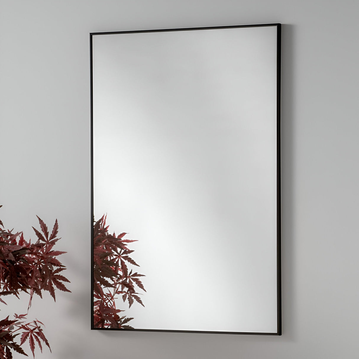 The Mirra Rectangle Wall Mirror - Black, featuring a sleek black frame and rectangular shape, is mounted on a plain white wall. The reflection of red foliage in the lower left corner enhances the minimalist home decor with a splash of color.
