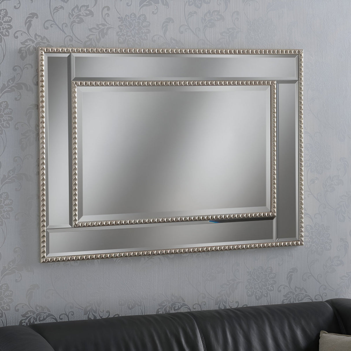 The Amalfi Rectangle Wall Mirror - 108x77cm, featuring its decorative beaded wood frame, is displayed on a patterned wallpapered wall above a dark leather couch. This luxurious mirror's ornate design enhances the room's decor with elegance.