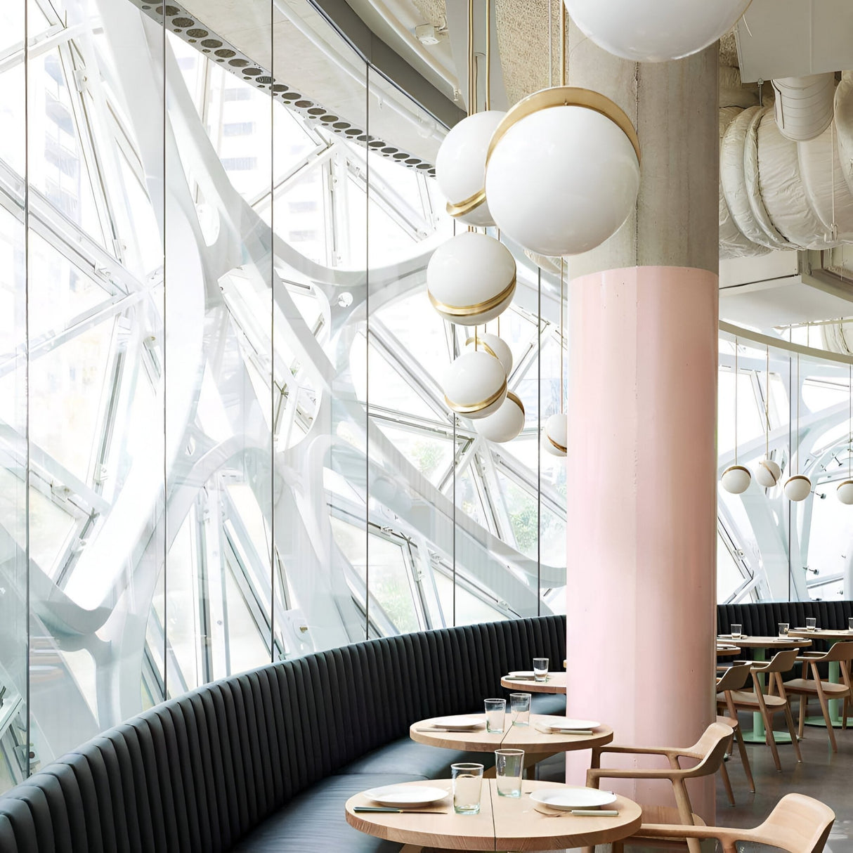 A modern restaurant with large glass windows, a curved dark bench, wooden tables set for dining, a pink-and-white column, and round Arc Pendant Lights in Gold & White creates a bright and airy atmosphere.