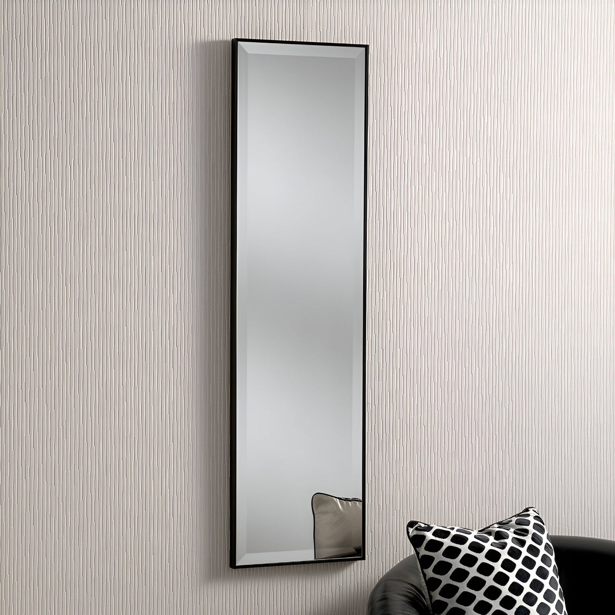 An Empyra Rectangle Wall Mirror in black hangs vertically on a textured cream wall, while a section of a black sofa with a patterned black and white pillow is visible beneath it.