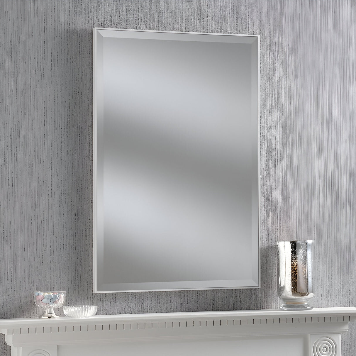 An Arden Rectangle Wall Mirror - White hangs on a textured, light gray wall above a white mantel. On the mantel are a small bowl and a tall glass vase, both featuring subtle decorative elements. The overall setting exudes simplicity and elegance.