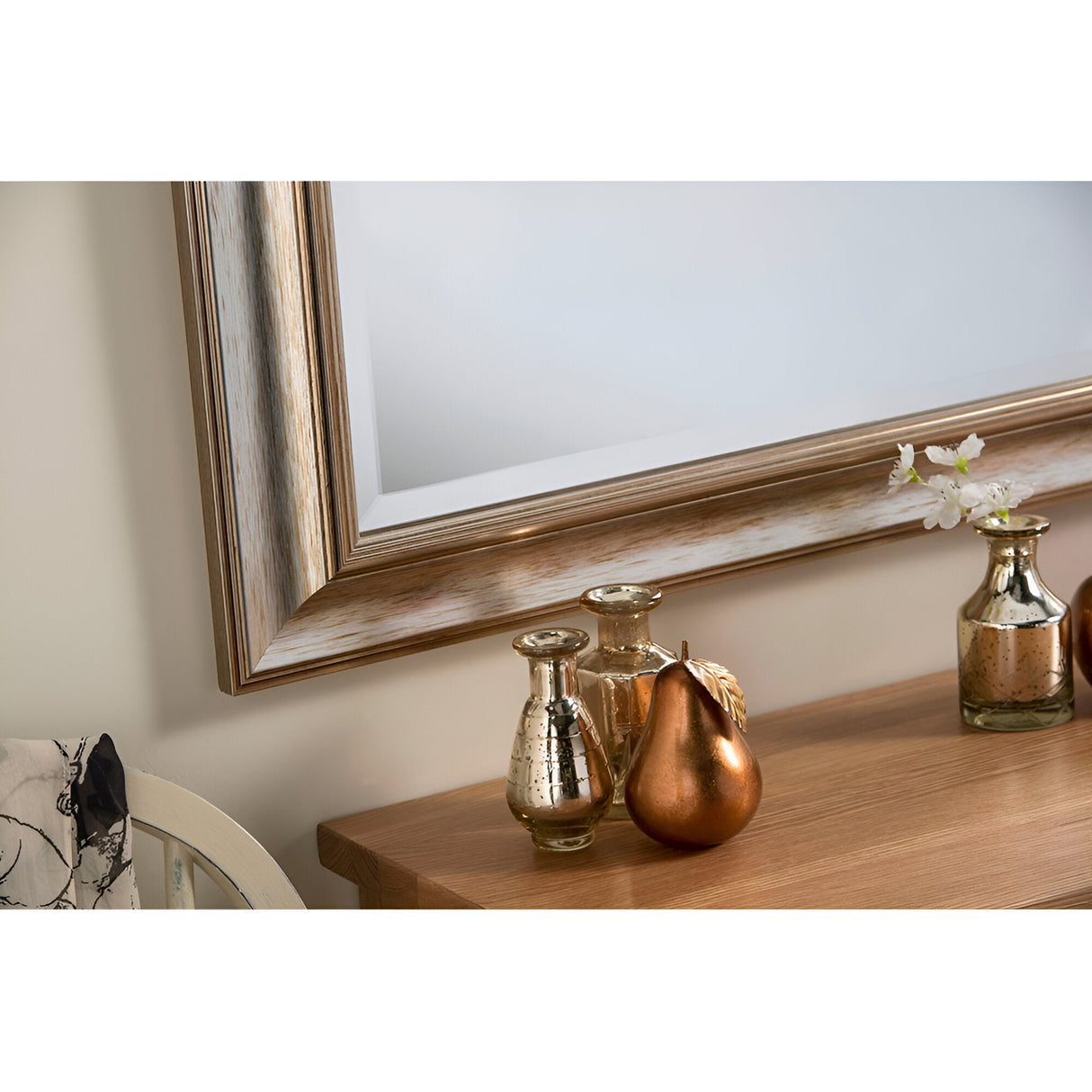 A wooden table beautifully decorated with home accessories, including metallic vases and a bronze pear-shaped ornament. Above it hangs the Aris Rectangle Wall Mirror with a sophisticated silver border that reflects light wonderfully. To the left, a small piece of fabric can be seen.