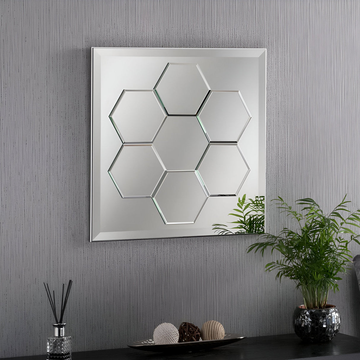 On a modern wall, an Arosa Rectangle Wall Mirror - 62x62cm is mounted on a textured gray surface. Below the mirror, a potted plant with green leaves is positioned next to a diffuser and decorative orbs, complementing the elegant touch of the sleek wood frame.