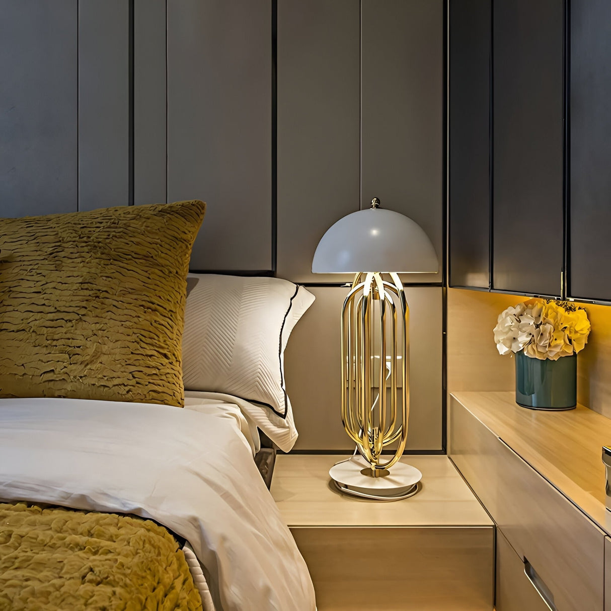 A modern nightstand topped with the Ascot 4 Light Large Table Lamp in white and gold, a small bouquet of yellow flowers, and a neatly made bed with white sheets and textured gold pillows against a dark paneled wall.