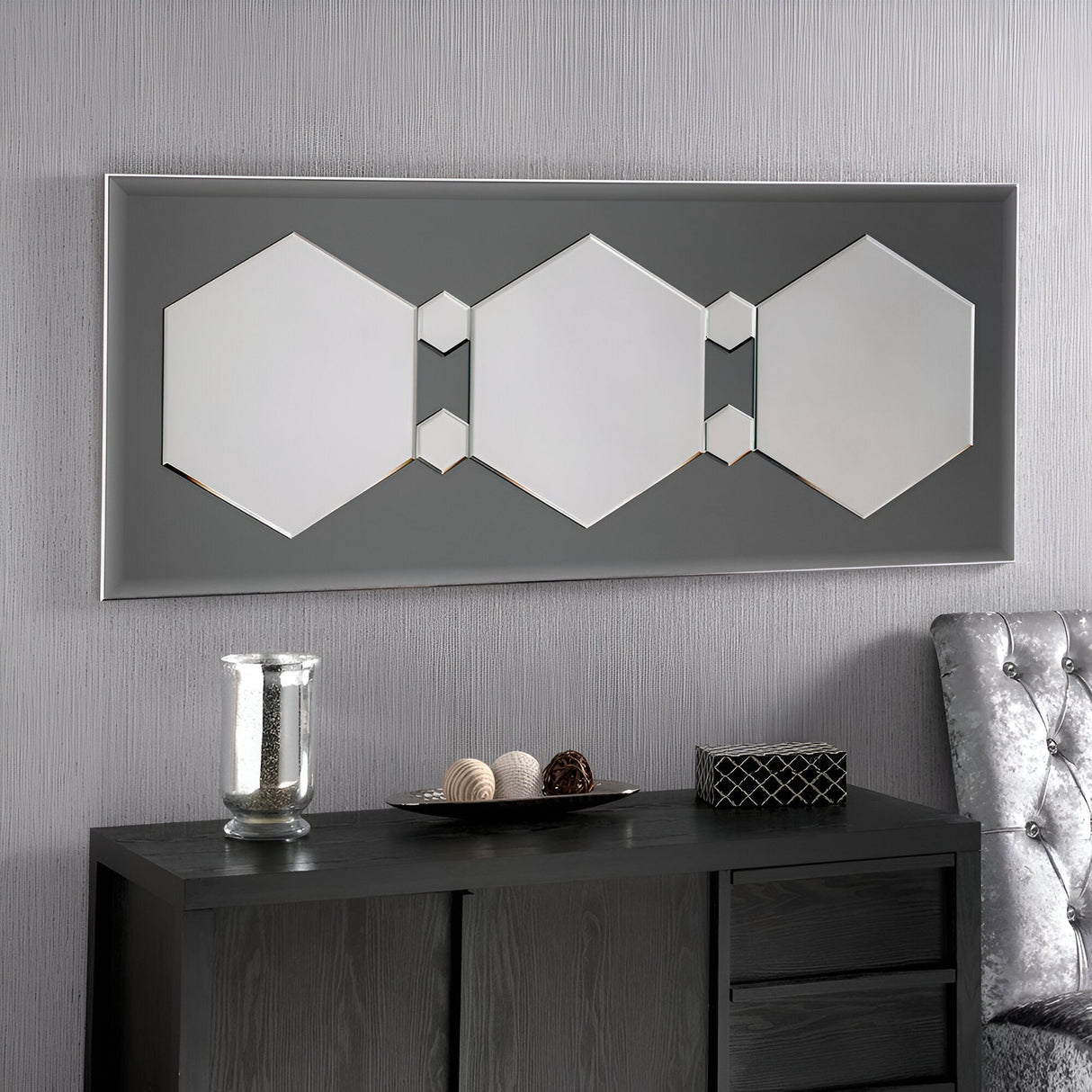 A modern room showcases the Athena Hexagon Wall Mirror in gray, measuring 154x62cm, mounted on a textured gray wall. Beneath it is a black console table adorned with a glass vase, decorative spheres, and a small box. A plush silver chair is partially visible on the right, adding to the contemporary decor.