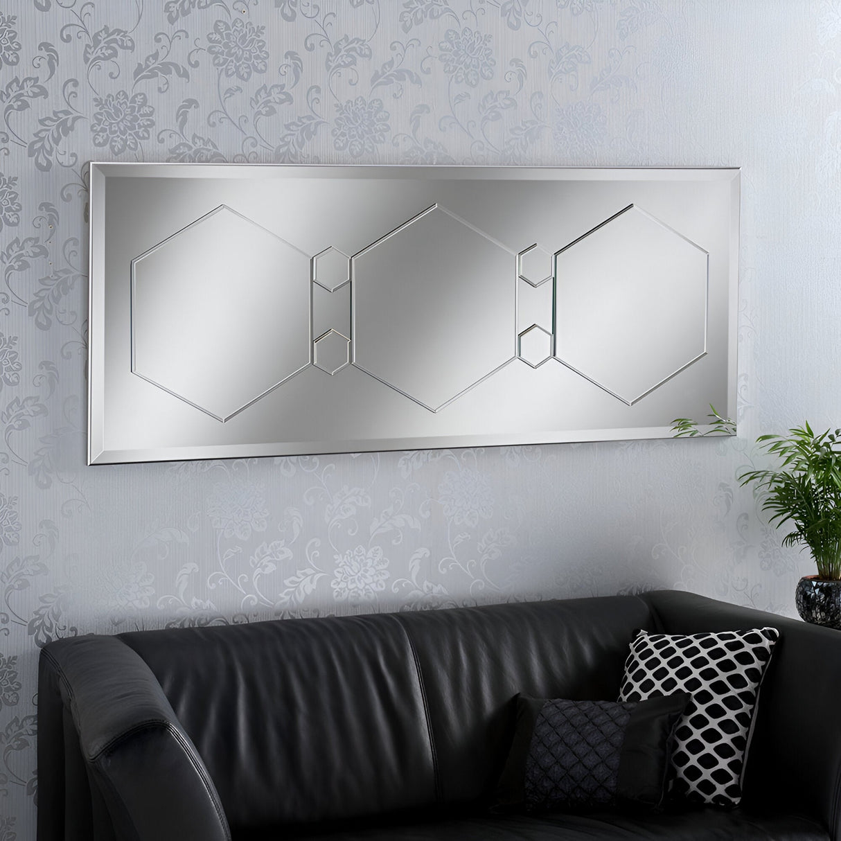A modern living room exudes contemporary elegance with the Athena Rectangle Wall Mirror Silver (154x62cm), showcasing a geometric design of hexagonal patterns above the black leather sofa. A patterned throw pillow adds charm, while a green potted plant complements the light floral wallpaper beautifully.