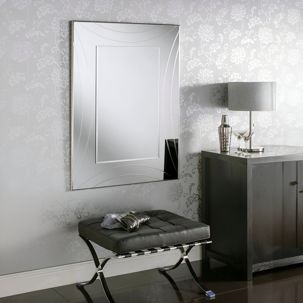 A chic interior features the Lumis Rectangle Wall Mirror Silver - 108x77cm on a patterned wall. Below, a tufted bench with a metallic finish rests on a glossy floor, exuding modern elegance. Completing the stylish look is a dark wooden cabinet with a sleek lamp and decorative items.