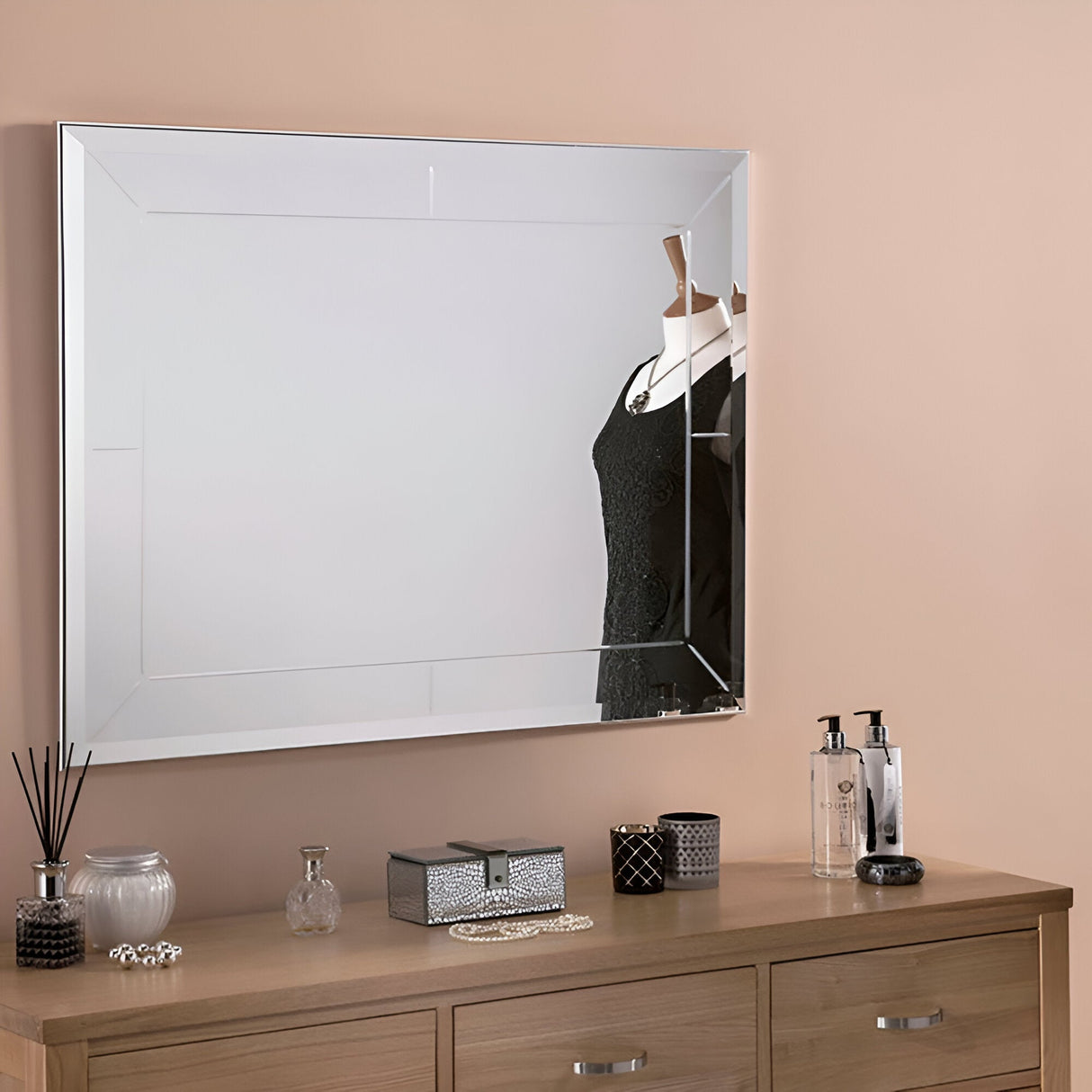 The Ray Rectangle Wall Mirror, placed against the beige wall, captures the reflection of a black lace dress displayed on a mannequin. Positioned beneath, a wooden dresser adorned with perfume bottles, a jewelry box, and various decorative items highlights its versatile design.