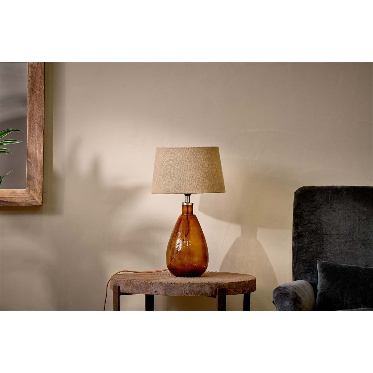 Baba Small Tall Recycled Glass Lamp Base in burnt amber with a teardrop body and linen shade, shown on a wooden side table in a living room setting.