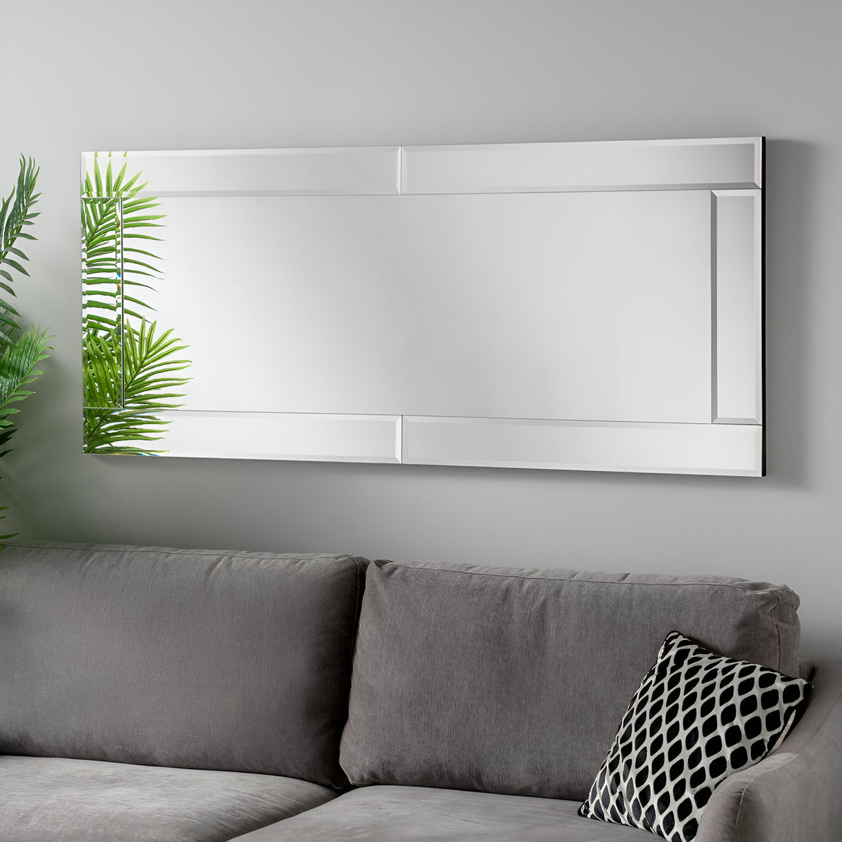 A Glim Rectangle Wall Mirror with a simple frame hangs on a light gray wall, embodying minimalist design. Below it, a gray sofa adorned with a black and white decorative pillow complements the contemporary home aesthetic. To the left, a green leafy plant adds a touch of nature.