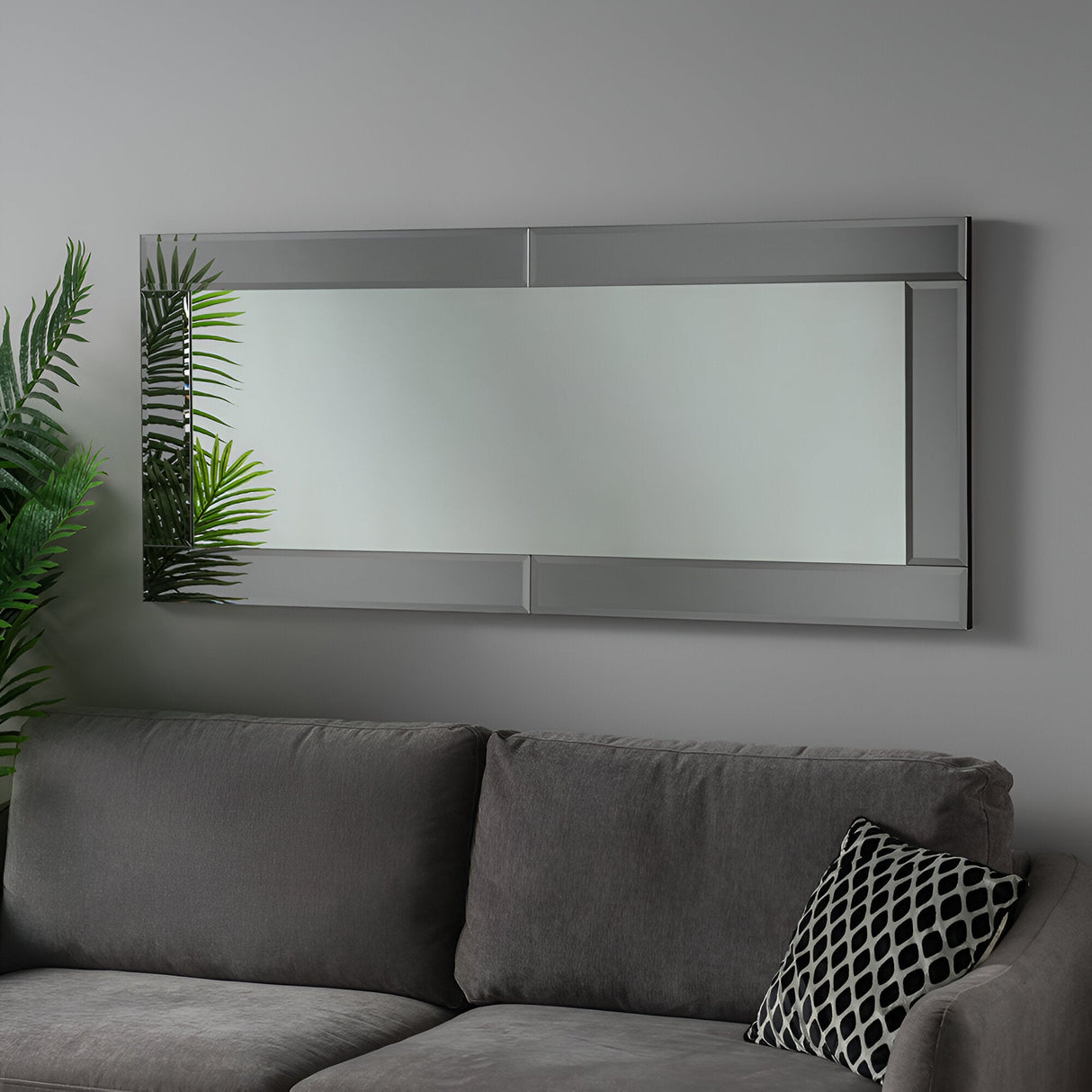 The Evocis Rectangle Wall Mirror - Grey hangs on a gray wall, reflecting the sleek modern interior with a plants partial reflection. Below it, a gray sofa is adorned with a stylish black and white patterned pillow.