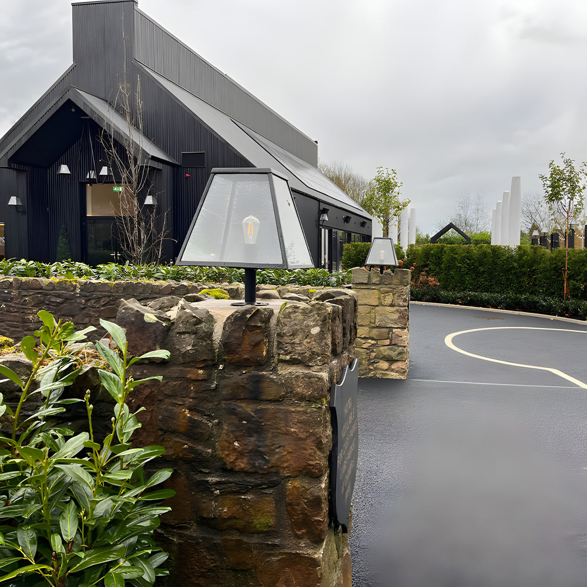 Large Contemporary Galvanised Steel Pillar Light in black, tapered glass-panelled design with exposed bulb, mounted on a stone gate pier outside a modern black-clad building.