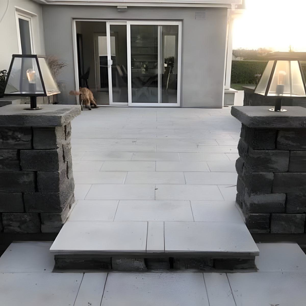 Small contemporary pillar light in black steel with tapered glass shade, mounted on a dark stone pier cap beside a patio and glass sliding door at dusk.