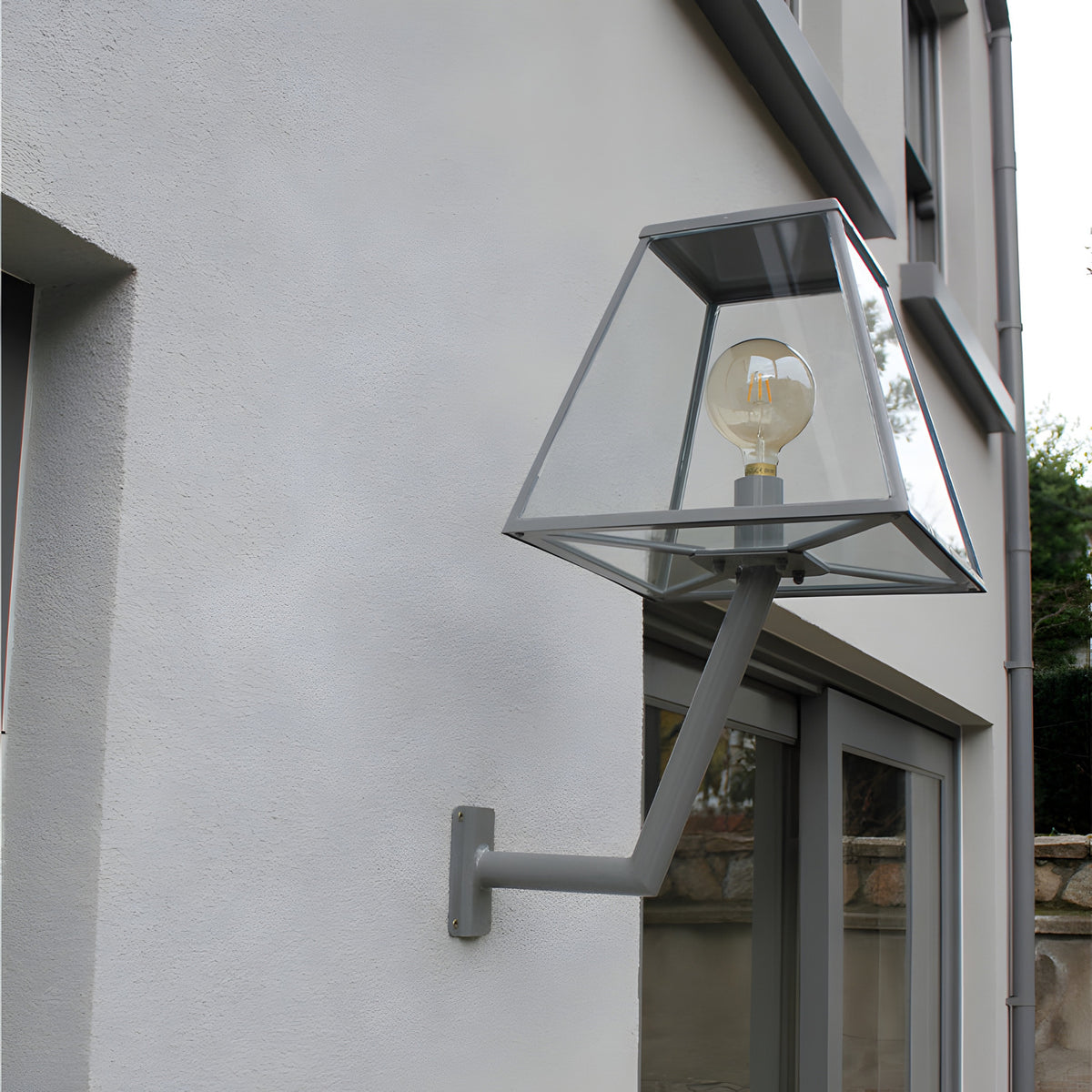 Large Contemporary Wall Lantern on an angled bracket in grey, mounted on an exterior wall beside a window, with a glass tapered shade and exposed globe bulb.