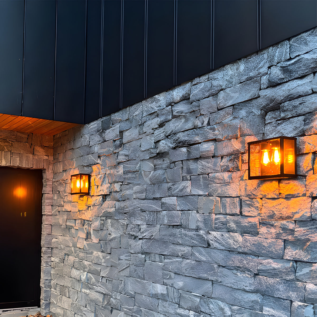 Contemporary Square Box Wall Lantern in black steel with glass panel, mounted on a stone wall beside a matching smaller lantern by a dark front door, warm bulb glow visible.