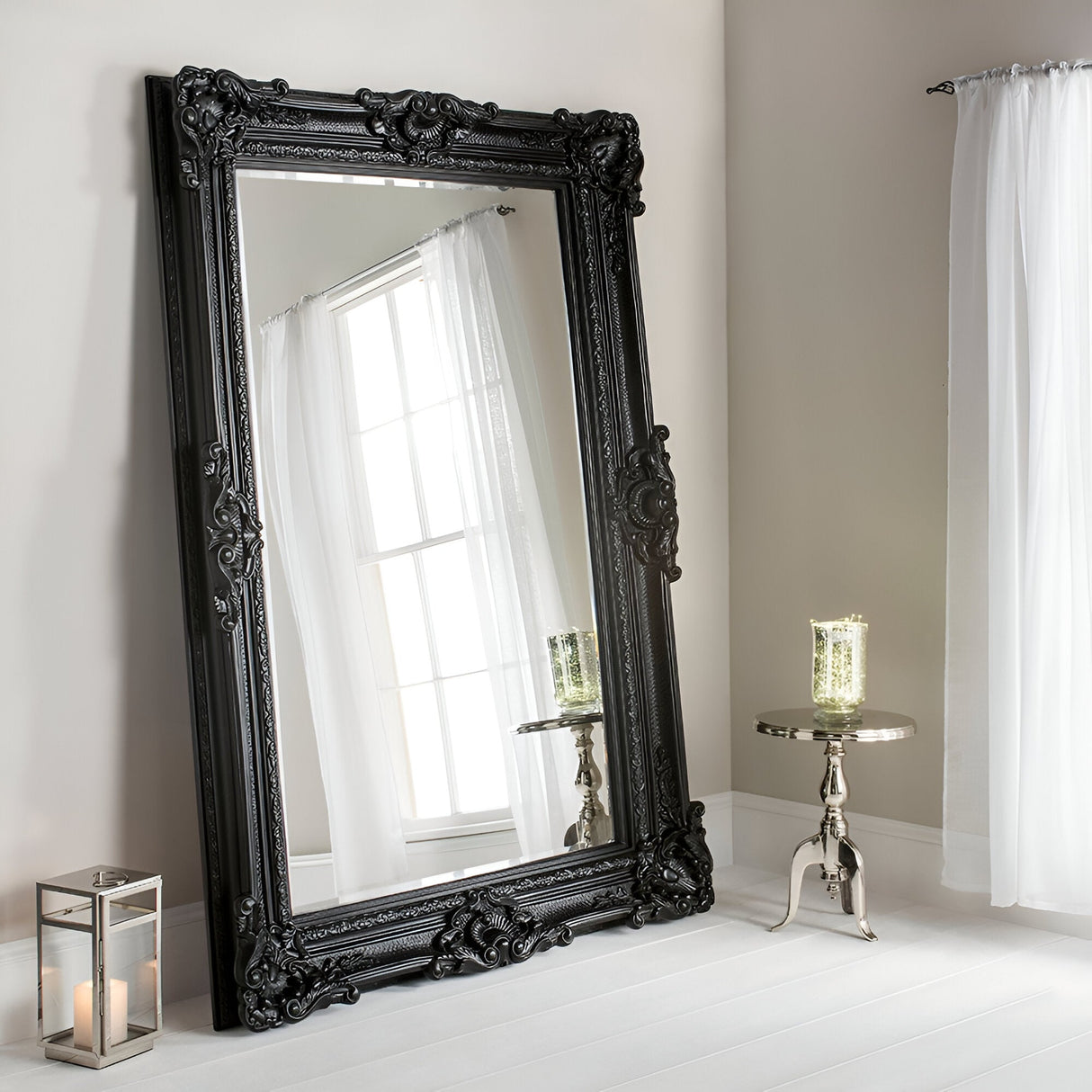 The Crystalix Leaner Mirror Black - 201x100 cm rests elegantly against a beige wall. Beside it, a small metallic table holds a lit candle in a decorative glass holder, while a lantern with candles sits gracefully on the floor. Sheer white curtains softly cover the nearby window.