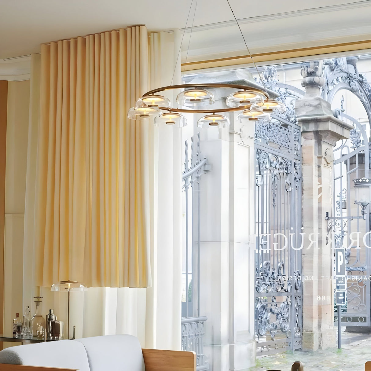 A modern dining area with beige curtains, a Blossom 6/8 LED Glass Chandelier in gold, a small bar setup, and large windows revealing ornate wrought iron gates outside.