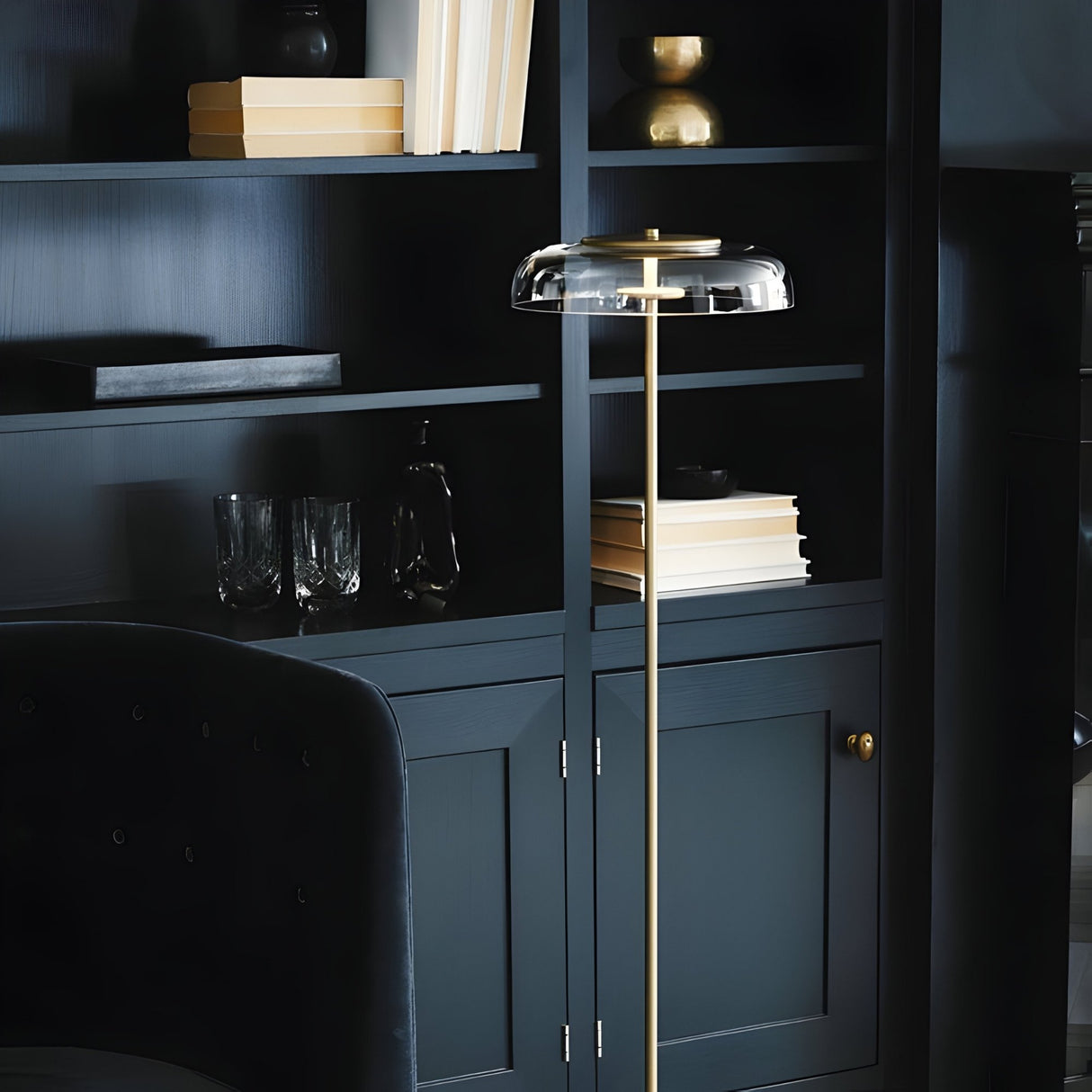 The Blossom LED Floor Lamp - Gold features a sleek gold stand and glass shade, illuminating dark built-in shelves filled with books, glassware, and decor in a stylish room while offering elegance and energy efficiency.