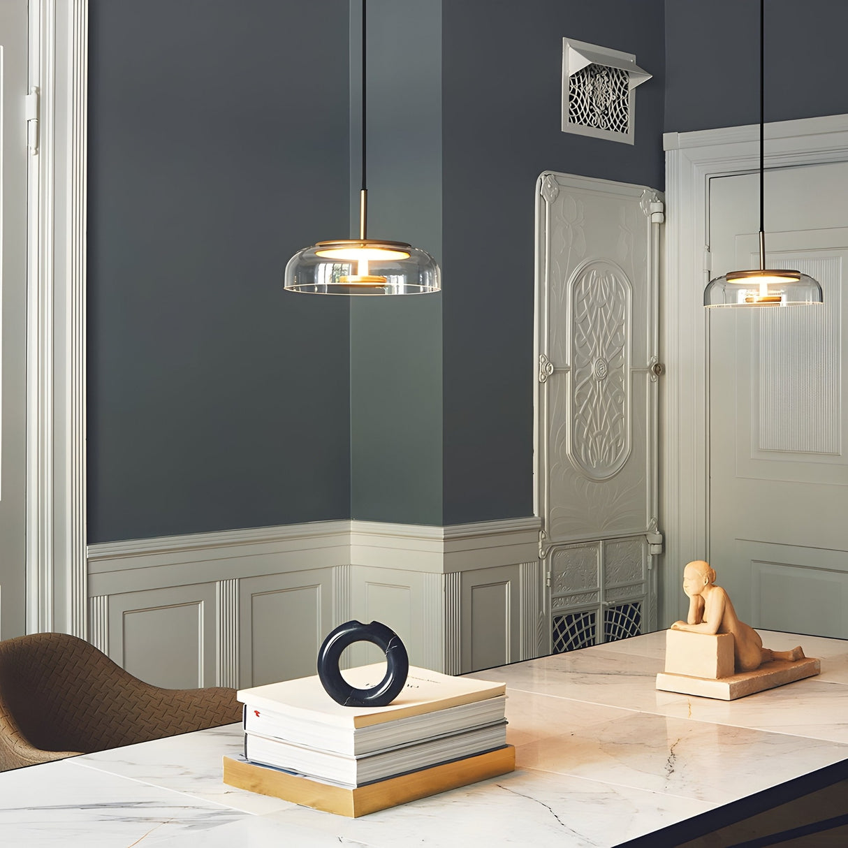 A modern interior features a marble table with stacked books, a circular sculpture, and a small figure. Overhead, the Blossom LED Glass Pendant Light - Gold brightens the space, enhanced by elegant molding and a vintage radiator cover.