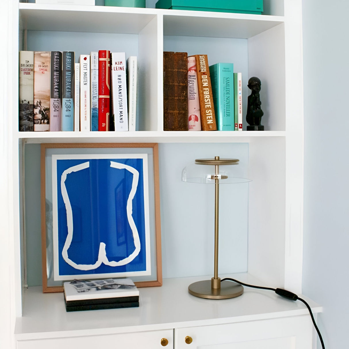 A white bookshelf holds books, a black sculpture, a framed blue and white print, two magazines, and a Blossom LED Glass Table Lamp - Gold with a handcrafted glass shade, adding contemporary style to the space.