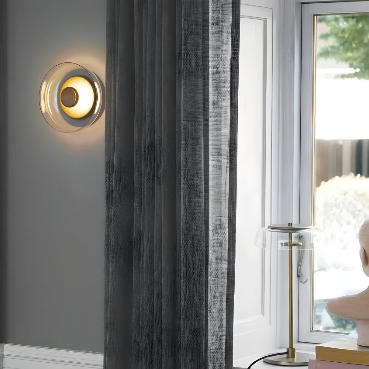 The Blossom LED Glass Wall & Ceiling Light - Gold, with its modern circular design, provides ambient lighting on a gray wall beside sheer gray curtains; nearby is a glass table lamp with a gold finish next to a decorative bust.