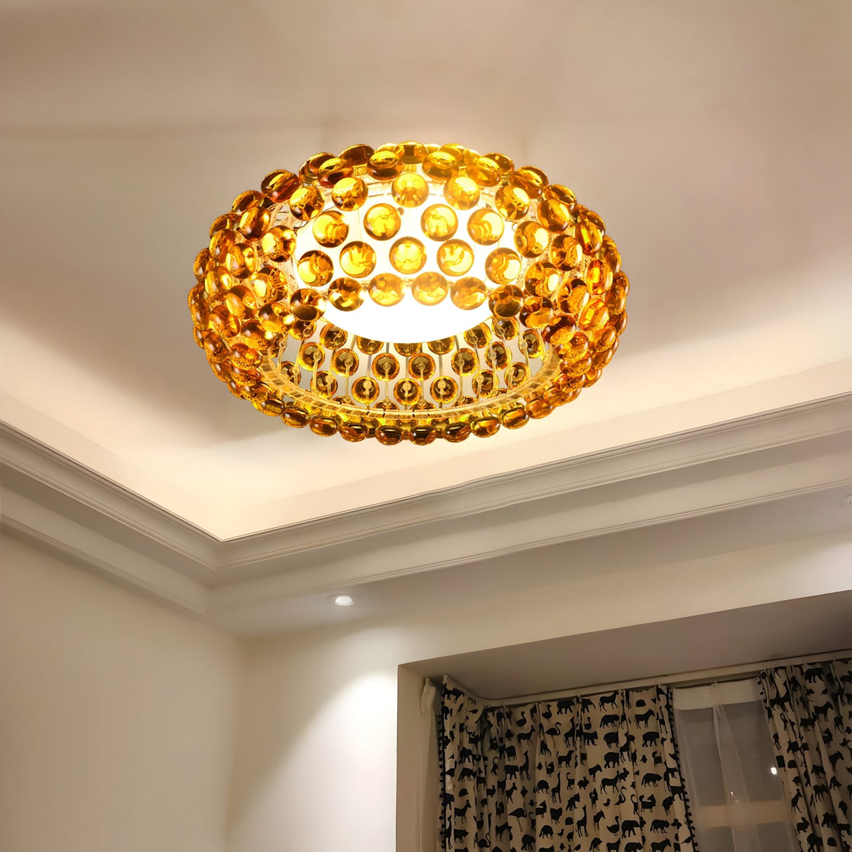 The Briller LED Ceiling Light in gold, featuring an amber, jewel-like design, brightens a room with white walls and curtains adorned with black animal silhouettes.