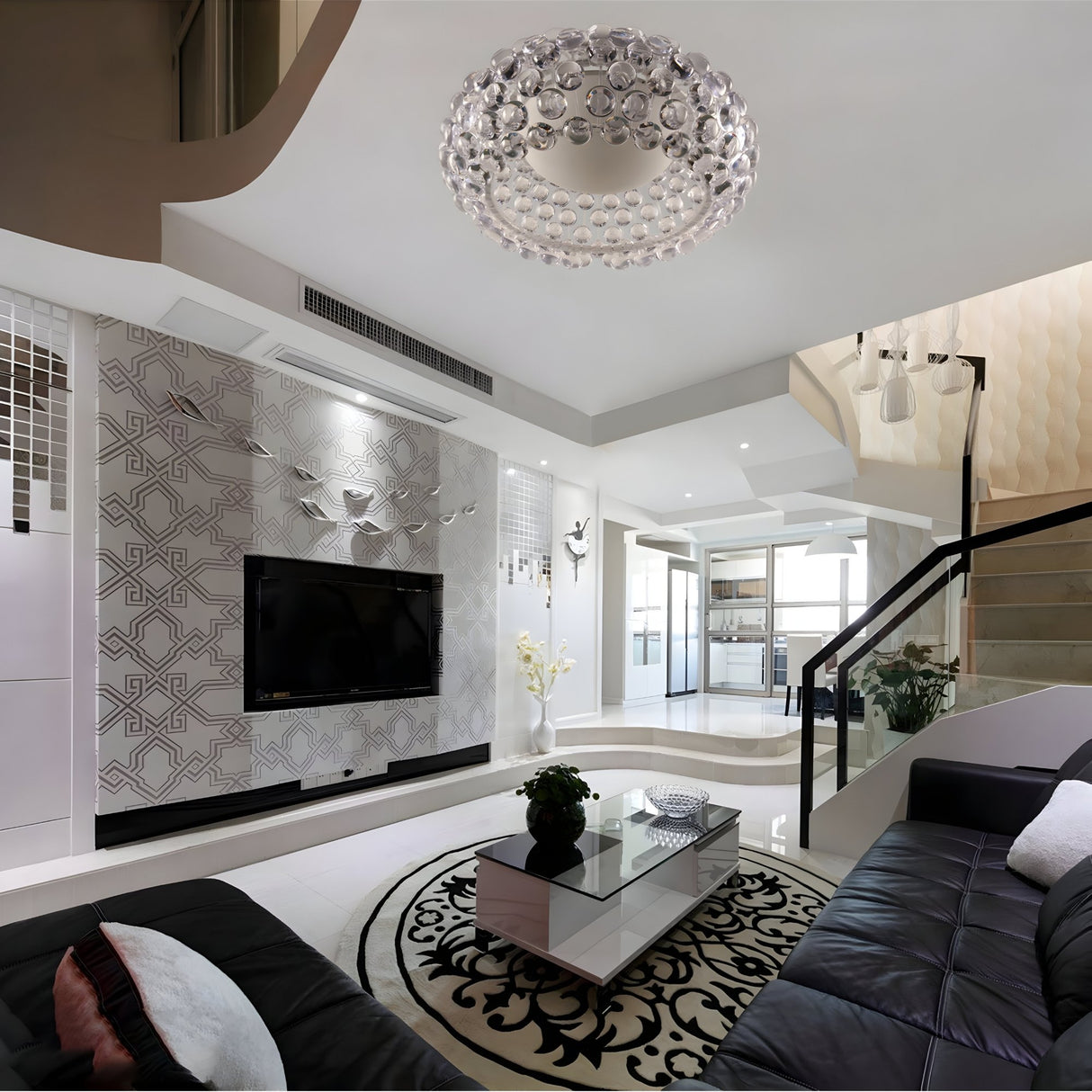 Modern living room with black leather sofas and glass coffee table on a patterned rug. The Briller LED Ceiling Light - Transparent adds a gem-like glow. TV on decorative wall, visible kitchen, stairs lead up; space feels bright and contemporary.
