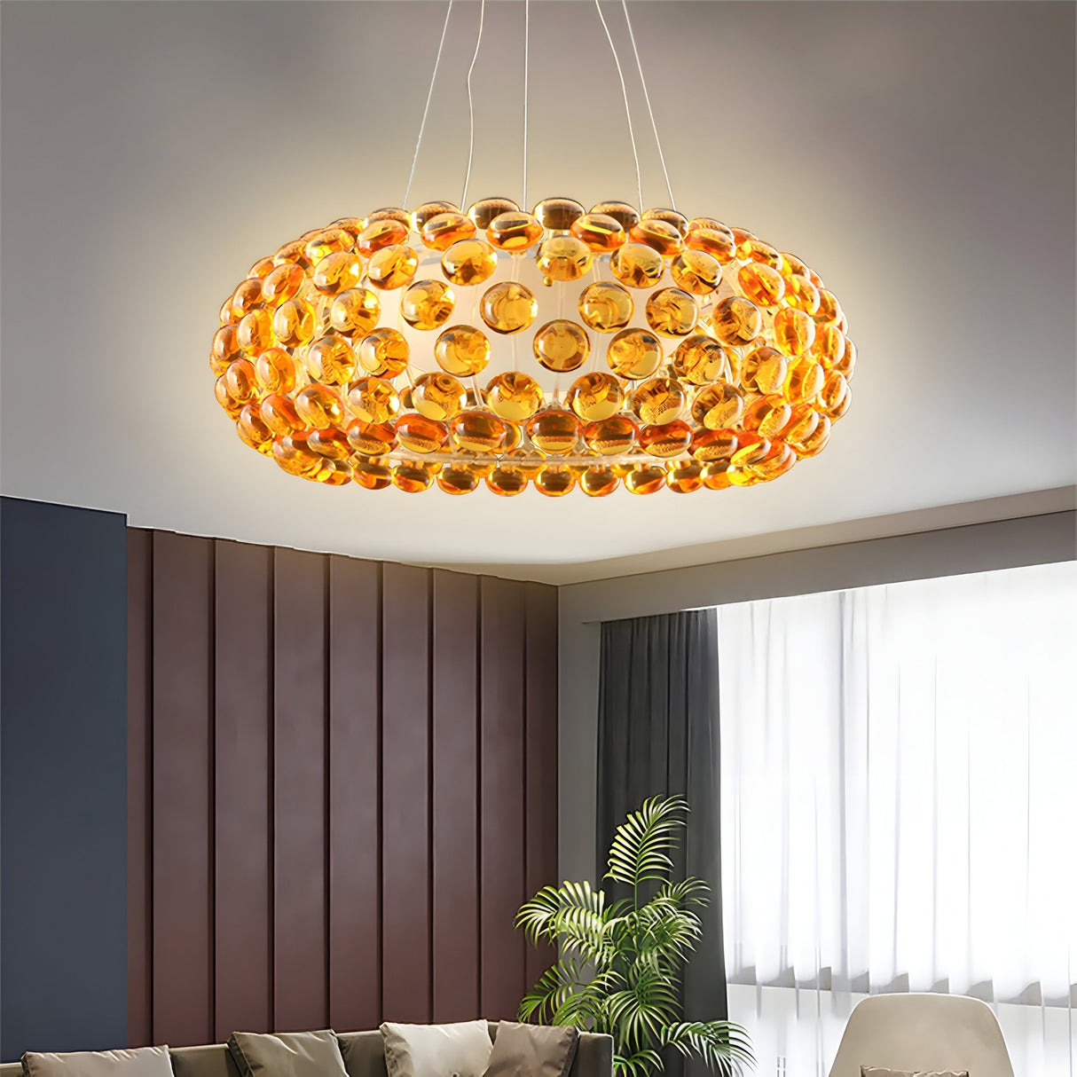 A modern living room features the Briller LED Large Suspension Light - Gold with amber glass spheres for ambient lighting. Below are a sofa and green plant by a window with sheer curtains, set against walls with vertical wood paneling.