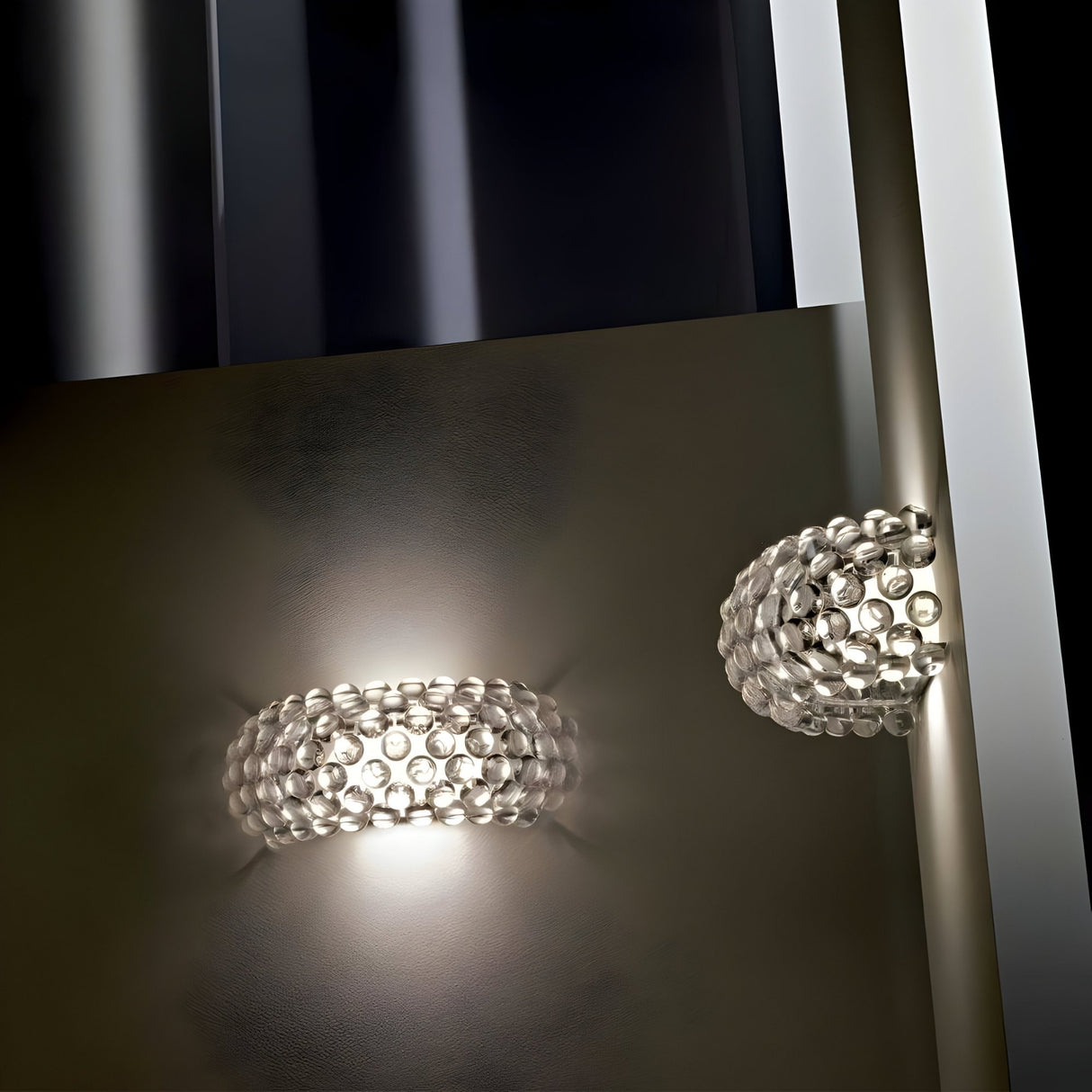 The Briller LED Large Wall Light in gold features a textured, bubble-like glass cover that emits a soft glow. Its reflection on a glossy surface enhances the modern effect with striking symmetry.