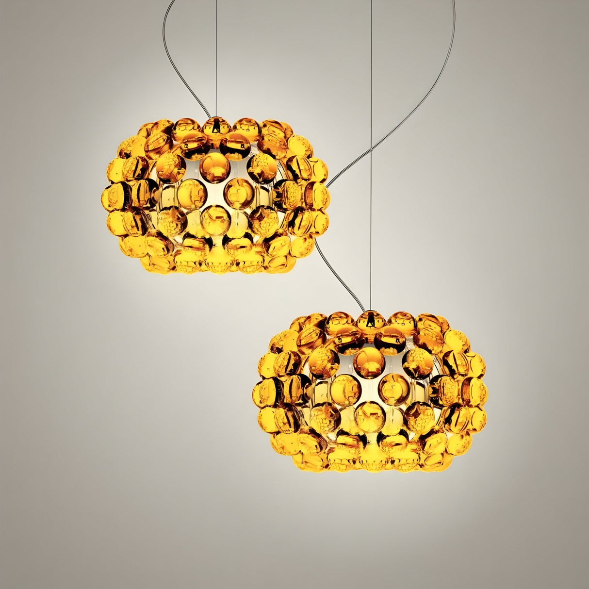Two Briller LED Small Pendant Lights in gold, each featuring a round amber acrylic sphere, hang by thin wires against a light background, casting a warm golden glow.