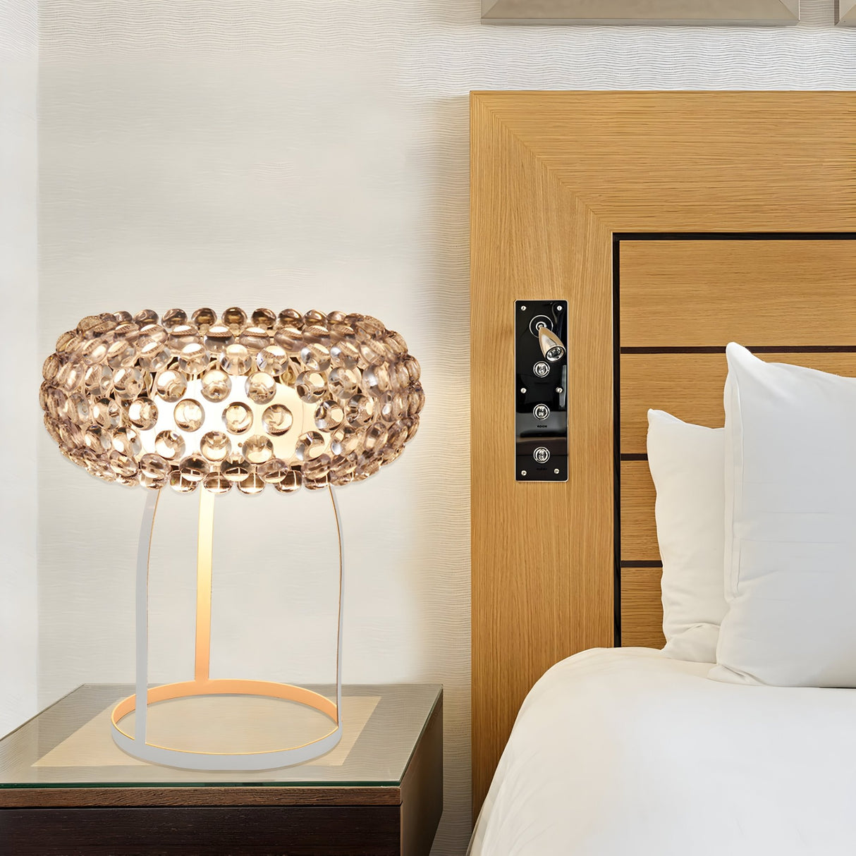 The Briller LED Table Lamp - Clear with a white base sits on a bedside table next to a wooden bed with white bedding, providing modern luxury and energy-efficient lighting for the bedroom.
