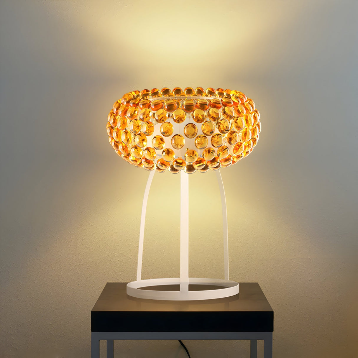 The Briller LED Table Lamp - Gold features a modern round shade with orange glass beads and a white metal stand, providing warm, energy-efficient light. Perfect on a dark square table against a light wall.