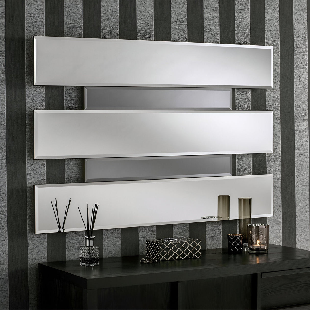 A modern wall features the Aiva Panel Wall Mirror measuring 122x81cm arranged in a sleek design against striped wallpaper. Below, a black console table enhances the contemporary decor with decorative items like a candle, a vase with reeds, and a textured box.