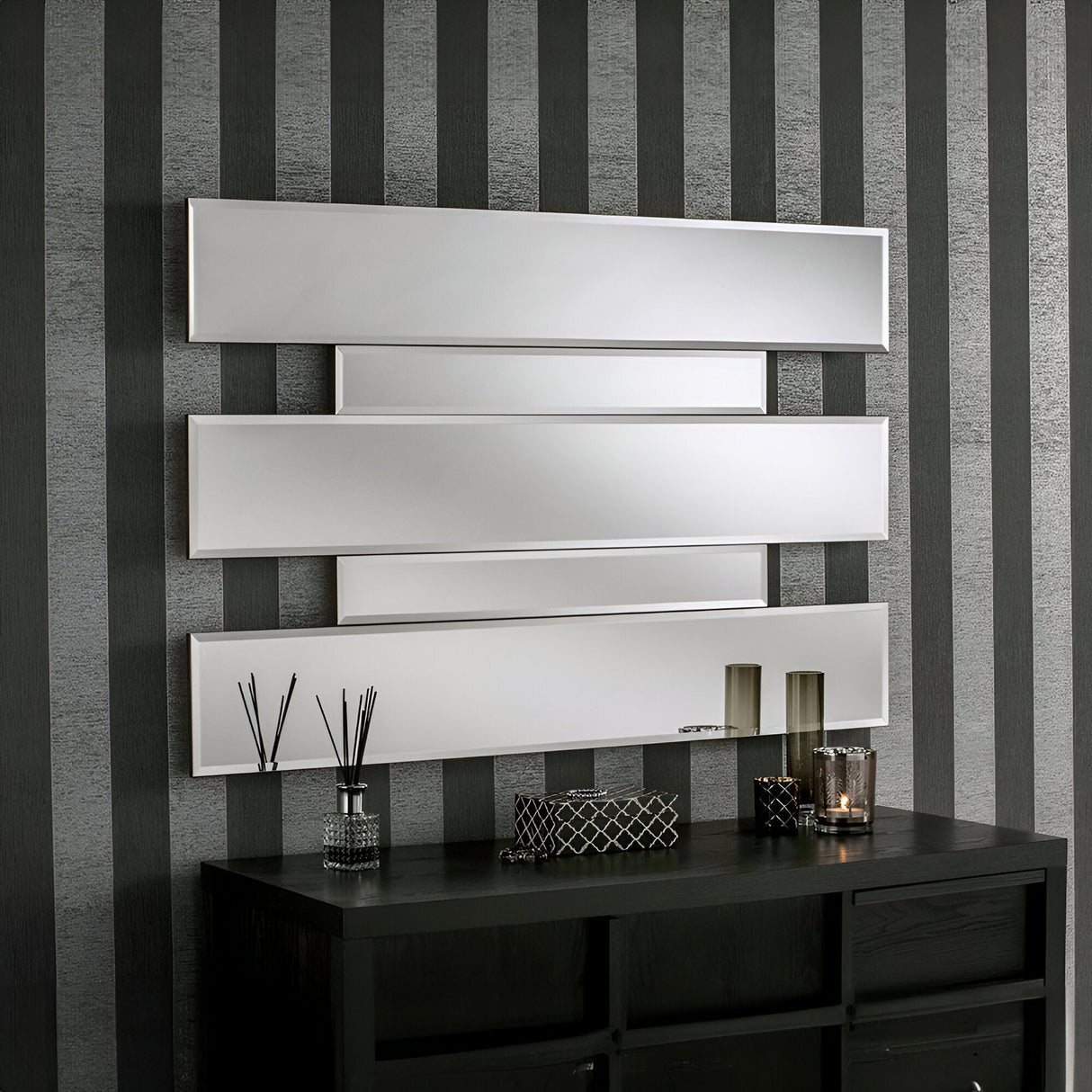 A minimalist interior features four horizontally aligned Aiva Panel Wall Mirrors in grey (122x81cm) on a striped gray and black wall. Below, a sleek black console table is adorned with carefully arranged decorative items such as candles and reed diffusers.
