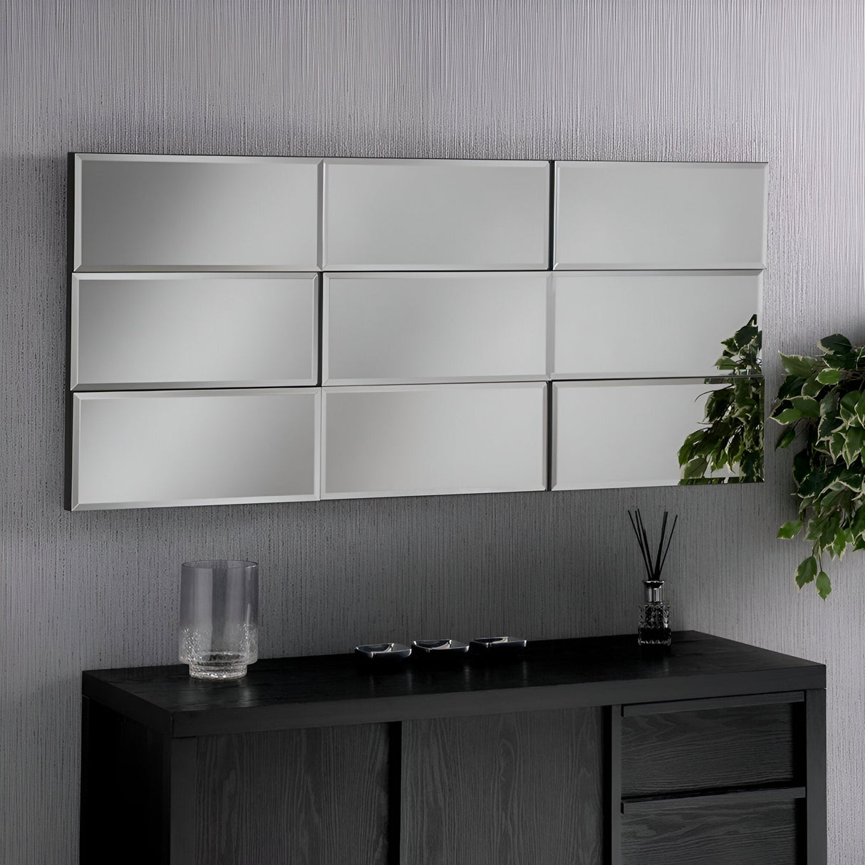 A modern room features a Luxia Rectangle Wall Mirror - 138x62cm, beautifully capturing contemporary design. Positioned horizontally above a dark wooden cabinet, the mirror accentuates three small decorative items and a clear glass vase. A leafy plant elegantly cascades from the side, adding a hint of greenery.