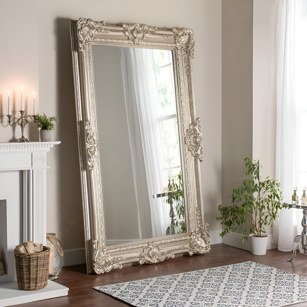 A Crystalix Leaner Mirror Silver - 201x100cm, featuring an ornate carved frame, leans against a beige wall. It reflects light from a bright window with sheer curtains. Close by, a potted plant brings greenery next to the patterned rug and a cozy fireplace adorned with candles and a wicker basket.