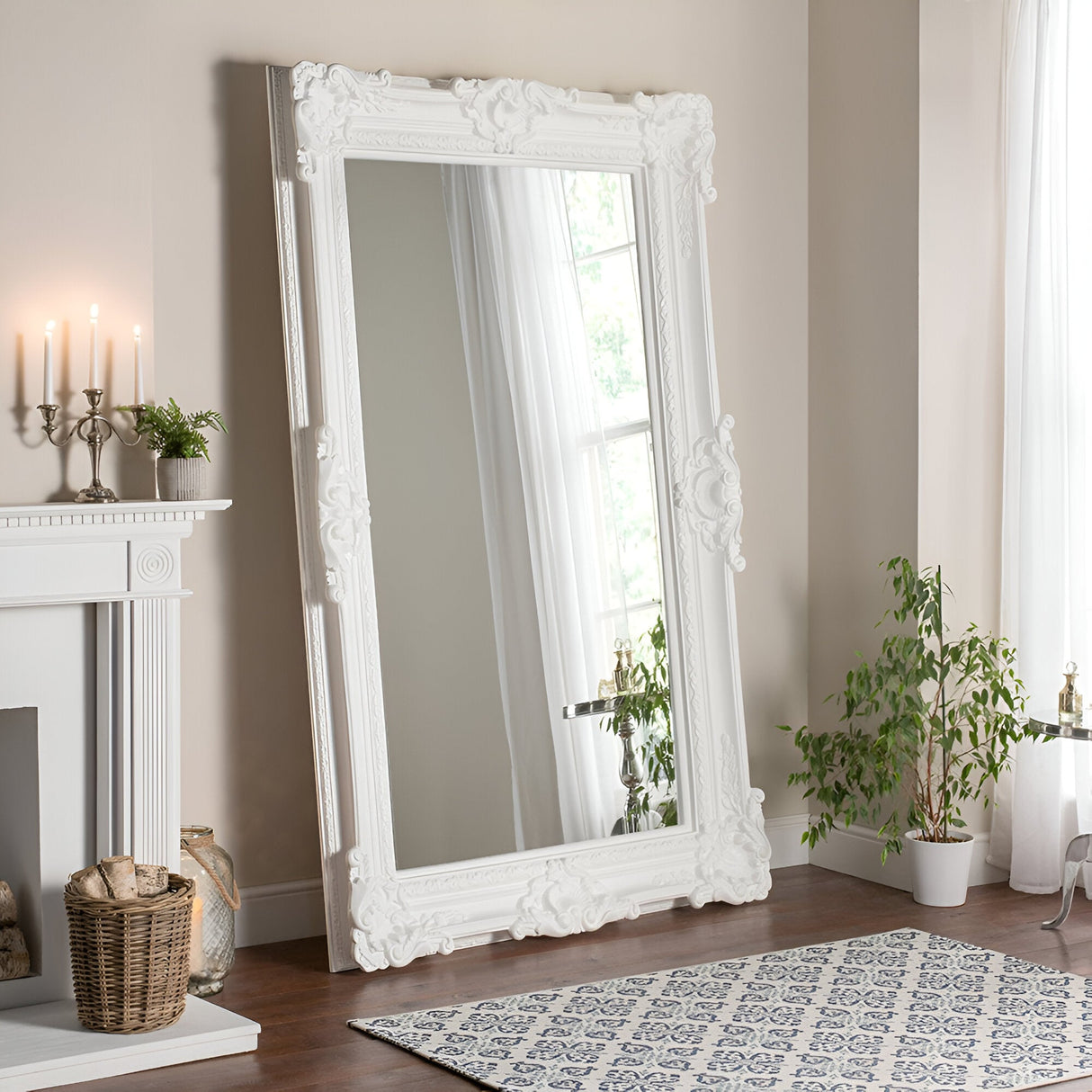 A sophisticated room showcases a sizable Crystalix Leaner Mirror White- 201x100cm, adorned with intricate details, resting against the wall and capturing the reflection of a lofty window draped with sheer curtains. A potted plant sits on a patterned rug nearby, while a decorated mantel featuring candles and a wicker basket adds to the ambiance.
