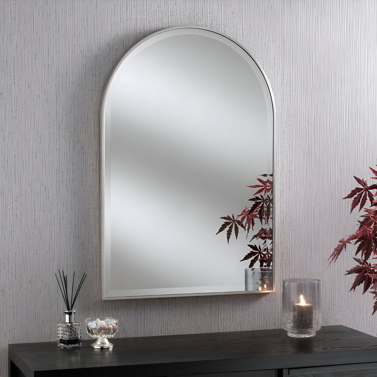 The Bellagio Arched Wall Mirror Silver - 93x63cm hangs on a textured gray wall above a black console table, which holds a glass vase with red leaves, a lit candle, a reed diffuser, and a small jar of candy.