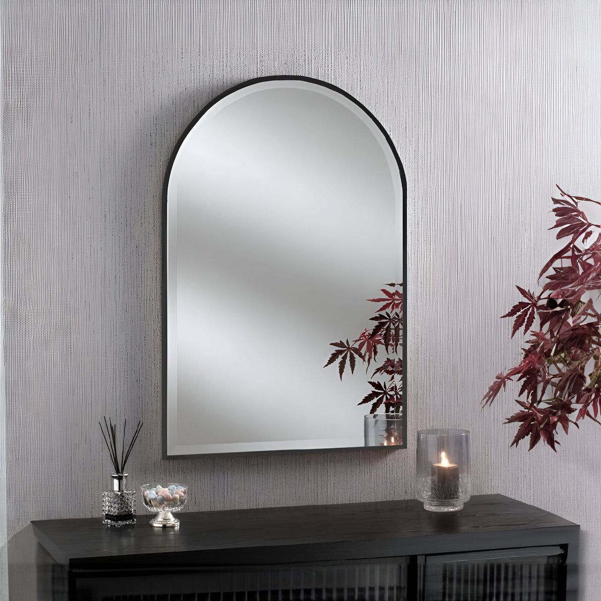 The Camden Arched Wall Mirror Black - 93x63cm, with its matte black finish, graces the patterned wall and reflects part of a plant with red leaves. Below, a minimalist interior setup features a glass candleholder with a lit candle, a jar of candies, and a reed diffuser on the sleek black table.