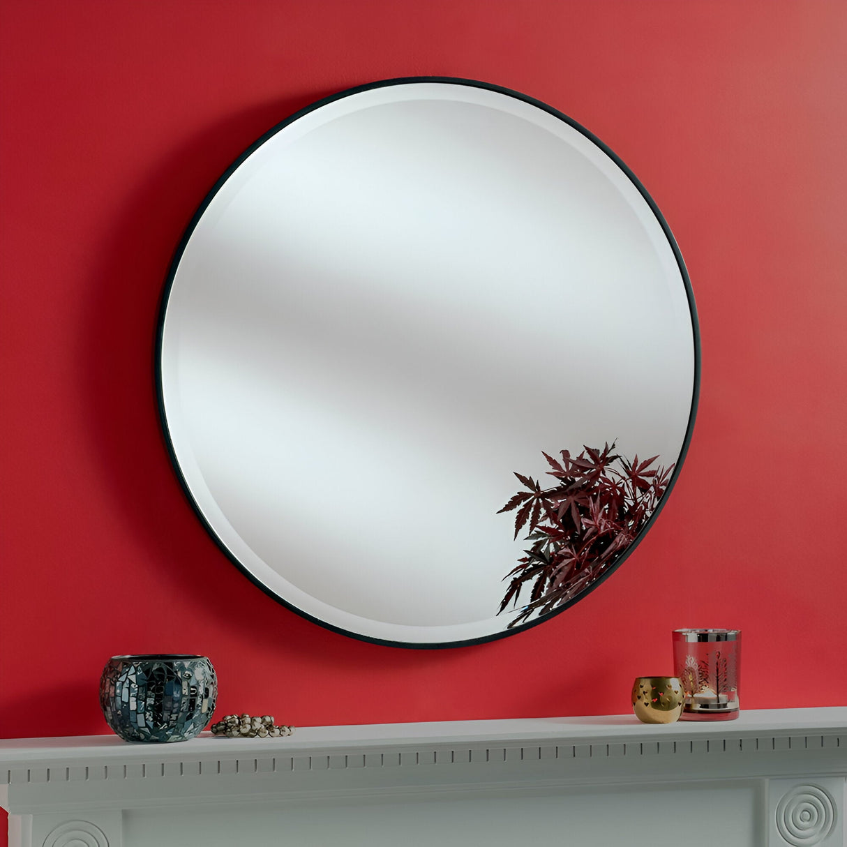 The Camden Circle Wall Mirror - Black, featuring a sleek circular design and black frame, is mounted on a red wall above a white mantel. The mirror partially reflects the leaves of a plant. Decorative items such as a mosaic-patterned vase and a candle holder adorn the mantel.