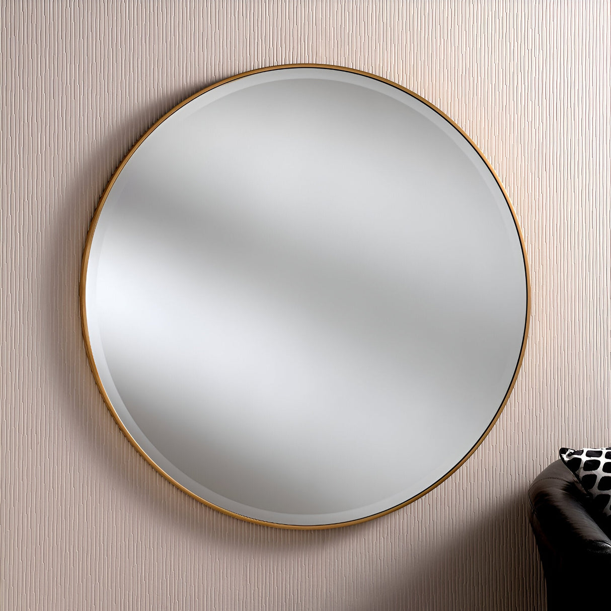 The Camden Circle Wall Mirror in gold, boasting a minimalist design, is mounted on a textured, light-colored wall. In the bottom right corner, a section of a black leather sofa adorned with a patterned cushion can be seen.
