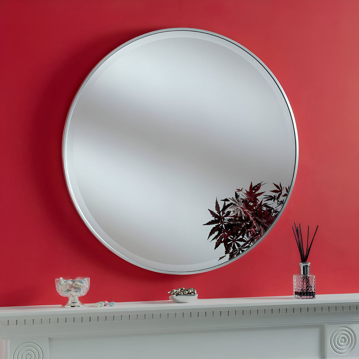 The Camden Circle Wall Mirror in silver, mounted on a red wall, reflects a small indoor plant. Below, the mantelpiece is adorned with home decor accents like a glass bowl and a decorative bottle with reed diffusers.