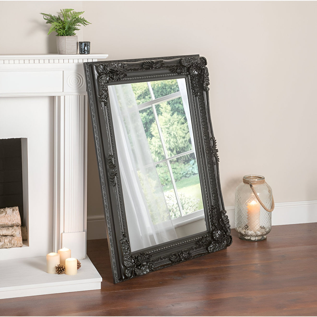 The Chantelle Rectangle Wall Mirror in black, measuring 119x89cm, leans against a white fireplace in the living room, exuding vintage charm. A lit candle rests inside a glass jar on the floor nearby, complemented by smaller candles and a green plant on the mantel.