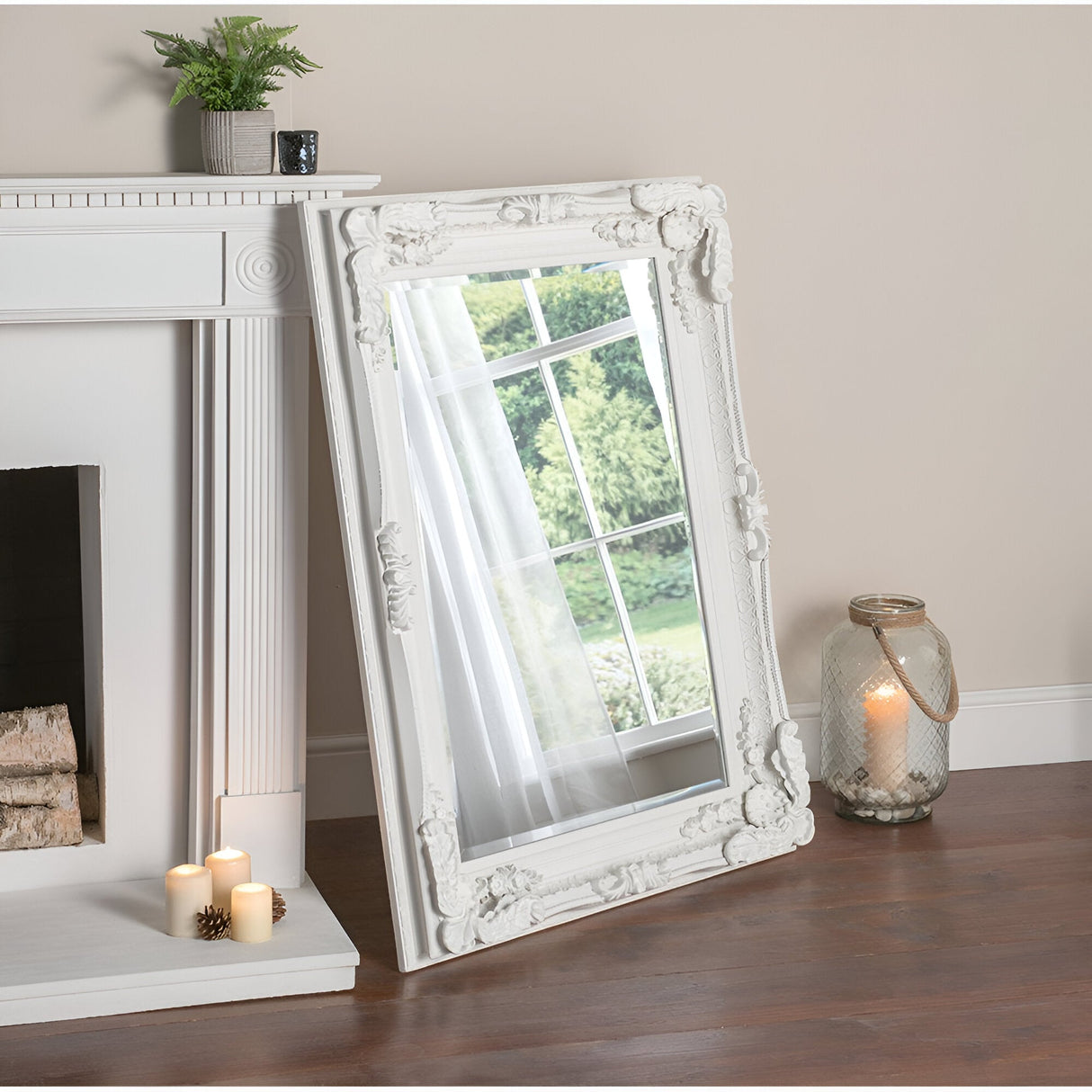 A Chantelle Rectangle Wall Mirror in white, measuring 119x89cm, leans elegantly against the wall, reflecting a window view of lush greenery. Nearby, the white fireplace adorned with lit candles and a glass lantern complements the wooden floor. A small plant rests gently on the mantel.