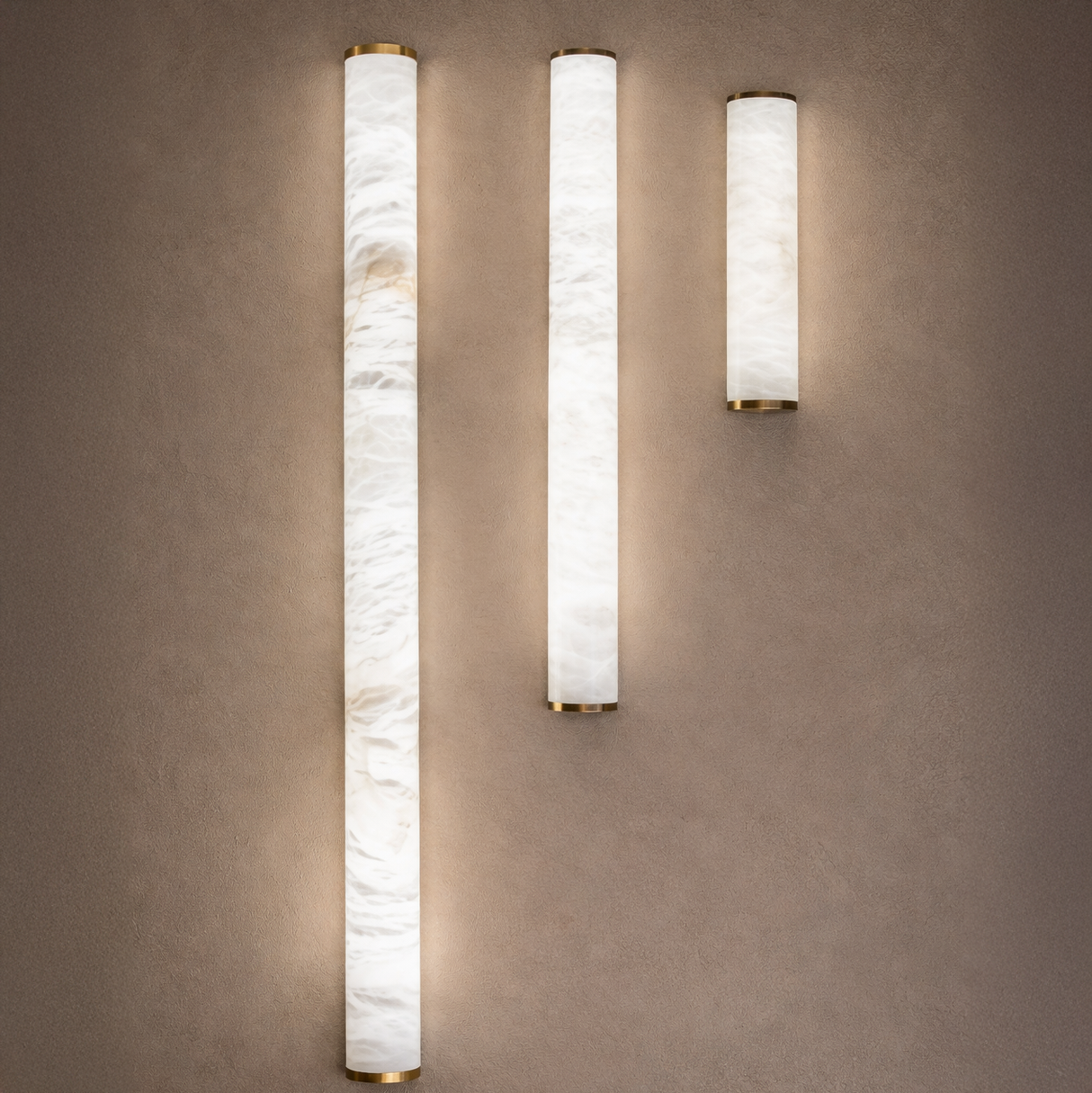 Three Corvane LED Large Slim Cylindrical Alabaster Wall Lights in soft white, featuring marbled alabaster and gold ends, are mounted vertically on a beige wall, casting a gentle, diffused glow.