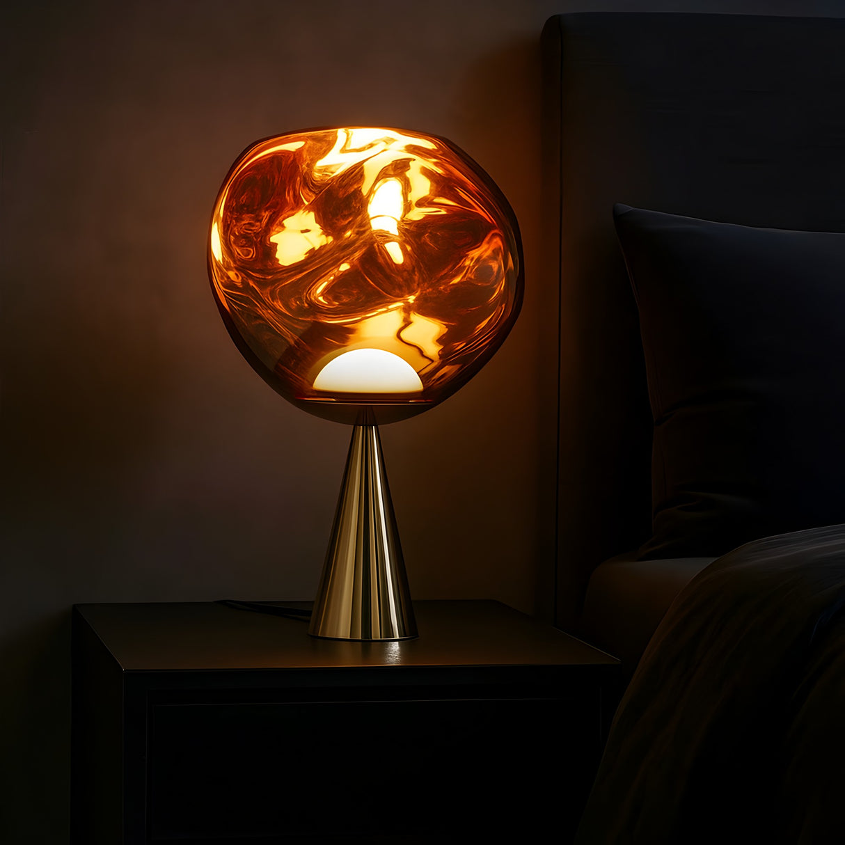 The Lava LED Table Lamp - Copper features a swirling amber glass shade that emits warm, golden light. Placed on a sleek black nightstand, it adds unique, modern lighting to any cozy, dimly lit bedroom.