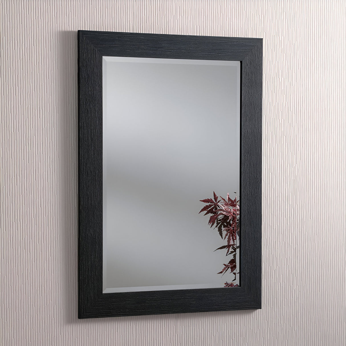 A Zennia Rectangle Wall Mirror - Black hangs on a textured light-colored wall, showcasing its minimalist reflection with part of a vibrant red-leaved plant in the bottom right corner.
